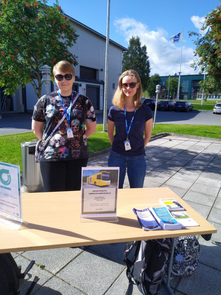 Kaksi henkilöä seisoo pöydän takana. Pöydällä on esitteitä.